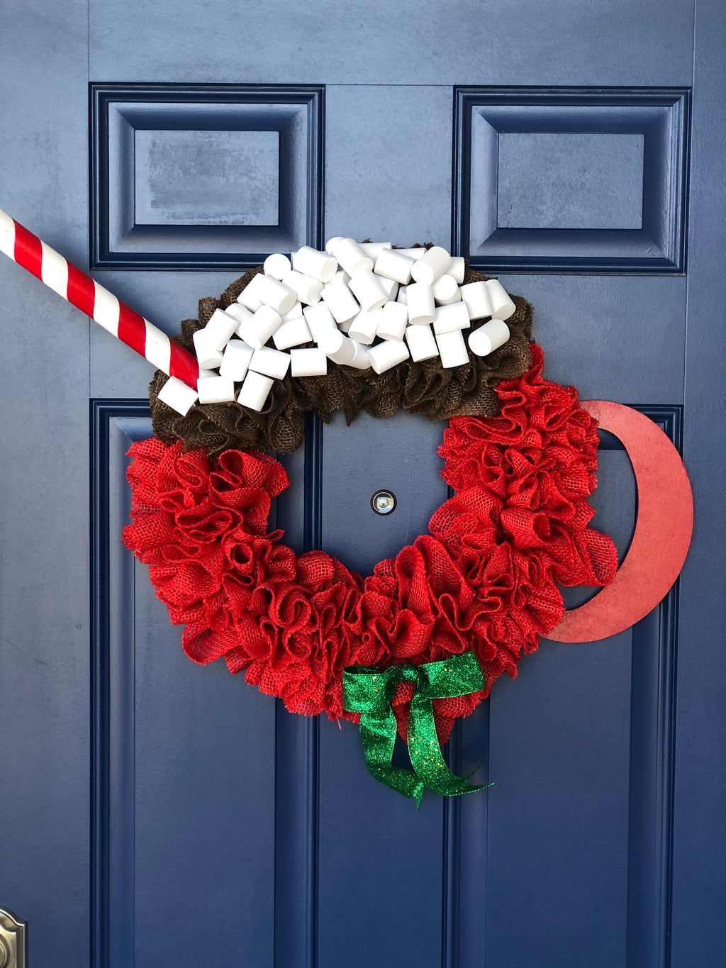 Red Burlap Wreath in the Shape of a Cup of Hot Cocoa with White Foam Marshmallows, a peppermint stick straw and a red wood handle on the cup with a green bow on a blue door. 