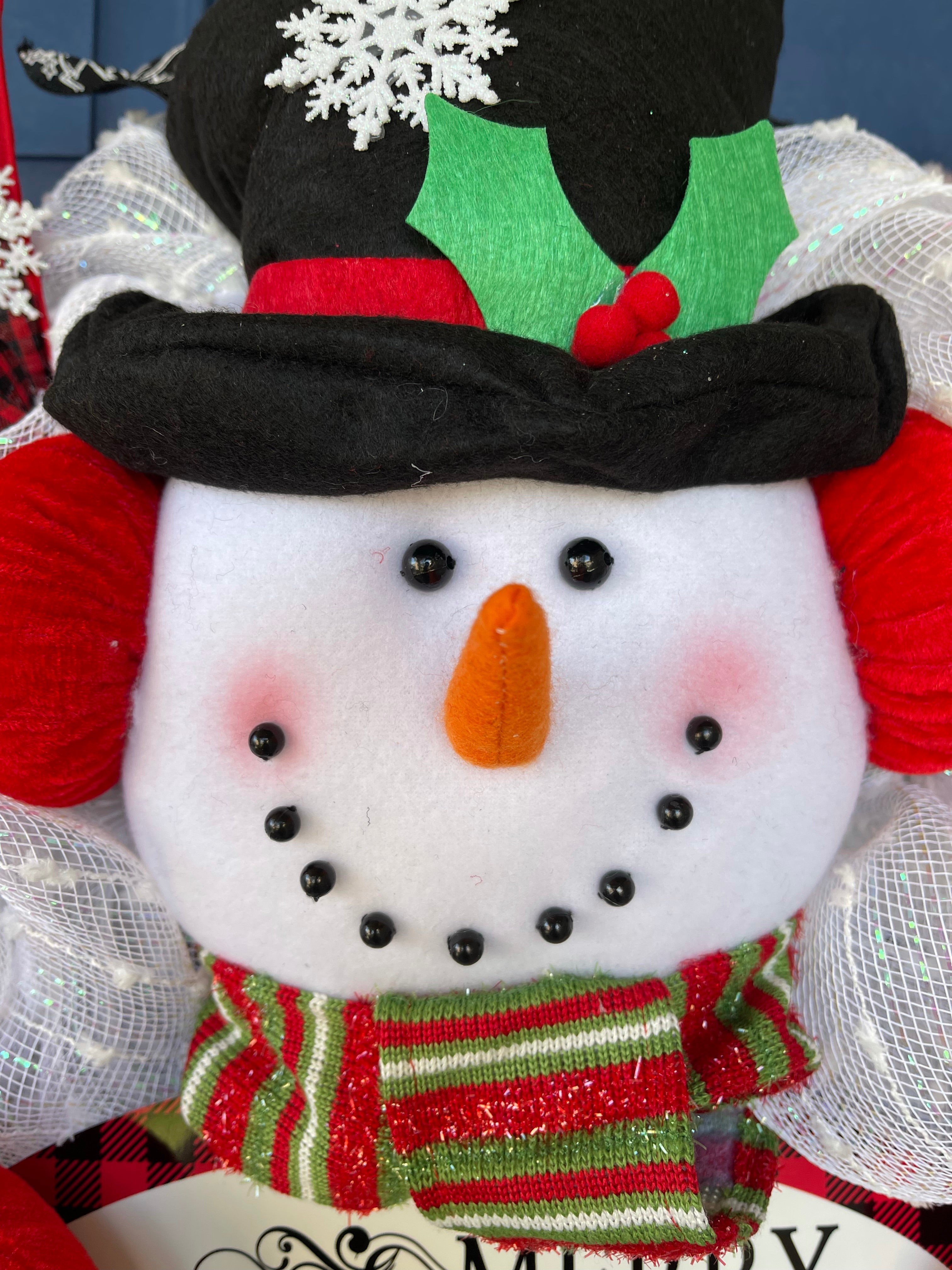 Close Up Detail of Plush Snowman Face with A Carrot Nose, wearing a Black Top Hat with a Snowflake, and Red Brim with Holly Berries, wearing Red Ear Muffs and a Green, White and Red Scarf