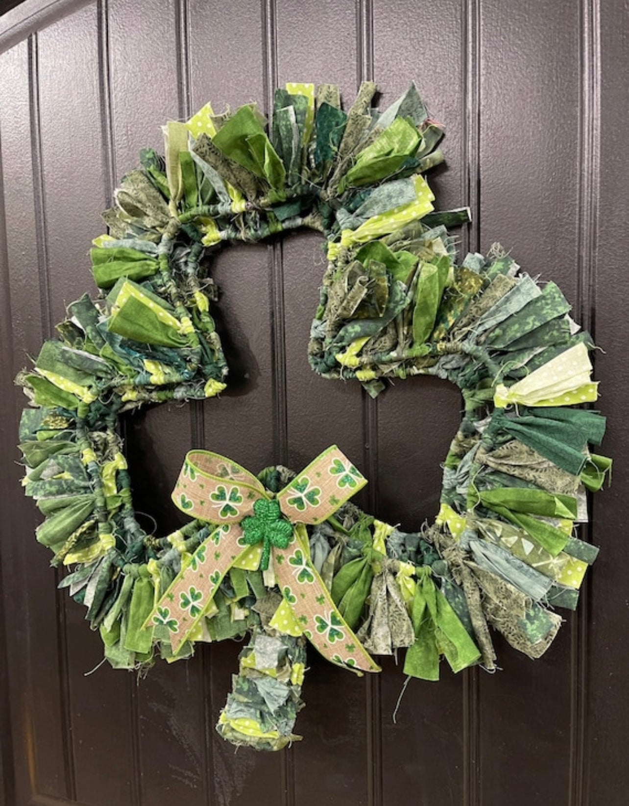 Right Side View of Various Light and Dark Shades of Green Cotton Fabric tied onto a Clover Shaped Frame topped off with a beige and clover ribbon and a glittered green shamrock on a black door. 