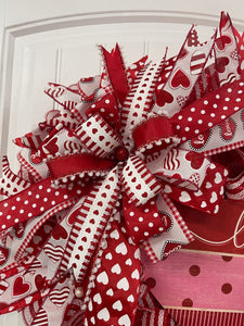Close Up Detail of Bow Featuring Ribbons with Red and White Hearts, Red Satin with White Tasseled Edges, Red with White Polka Dots, White Hearts on a Red Ribbon, and Lots of Hearts on White with a Red Glitter Ball in the Center. 