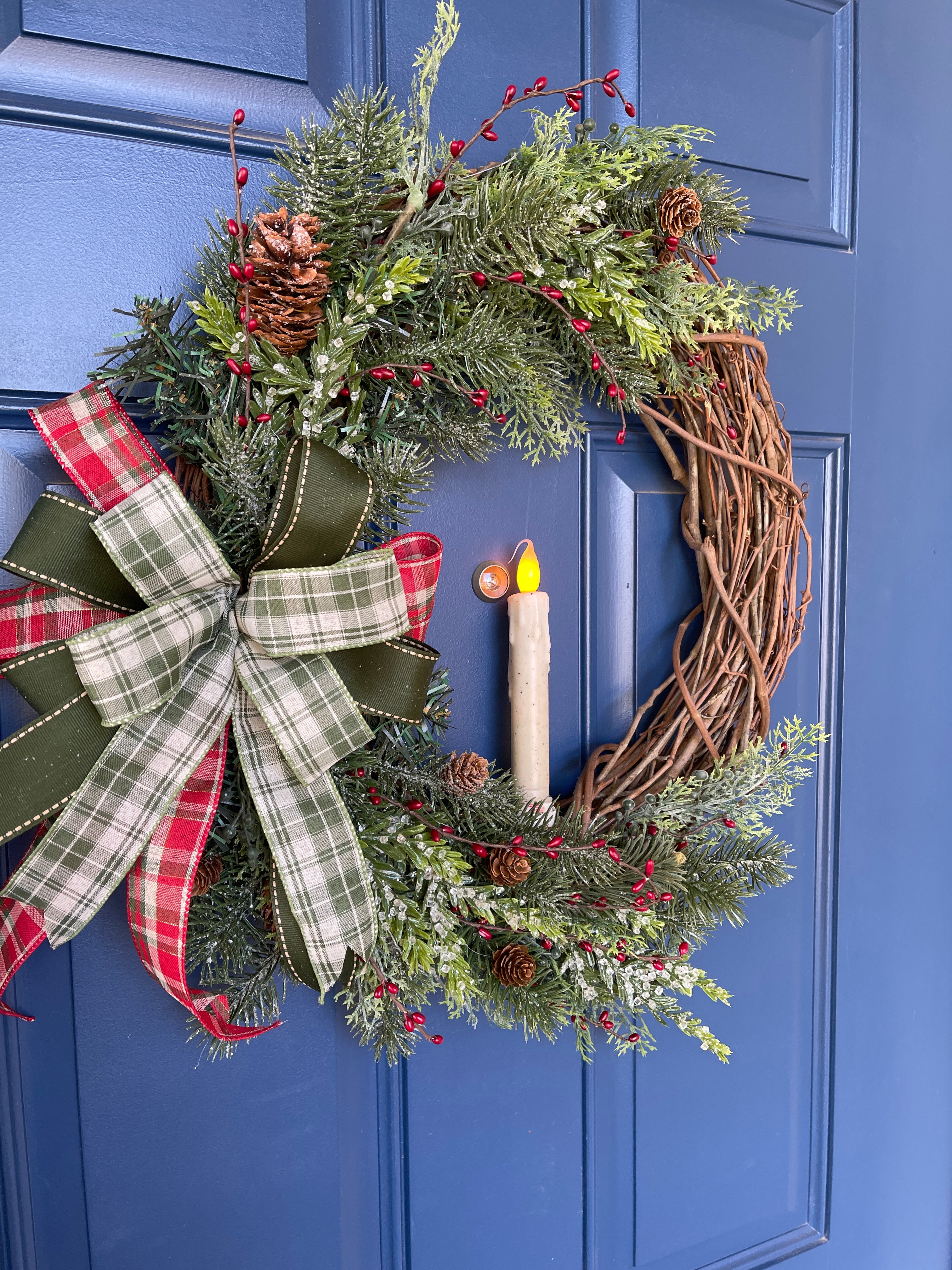 Left Side View of Christmas Grapevine Wreath with Artificial Pine Branches, Red Berries, Pinecones, with a Red, White and Green Bow and an Artificial Battery Operated Candle in the Center on a Blue Door