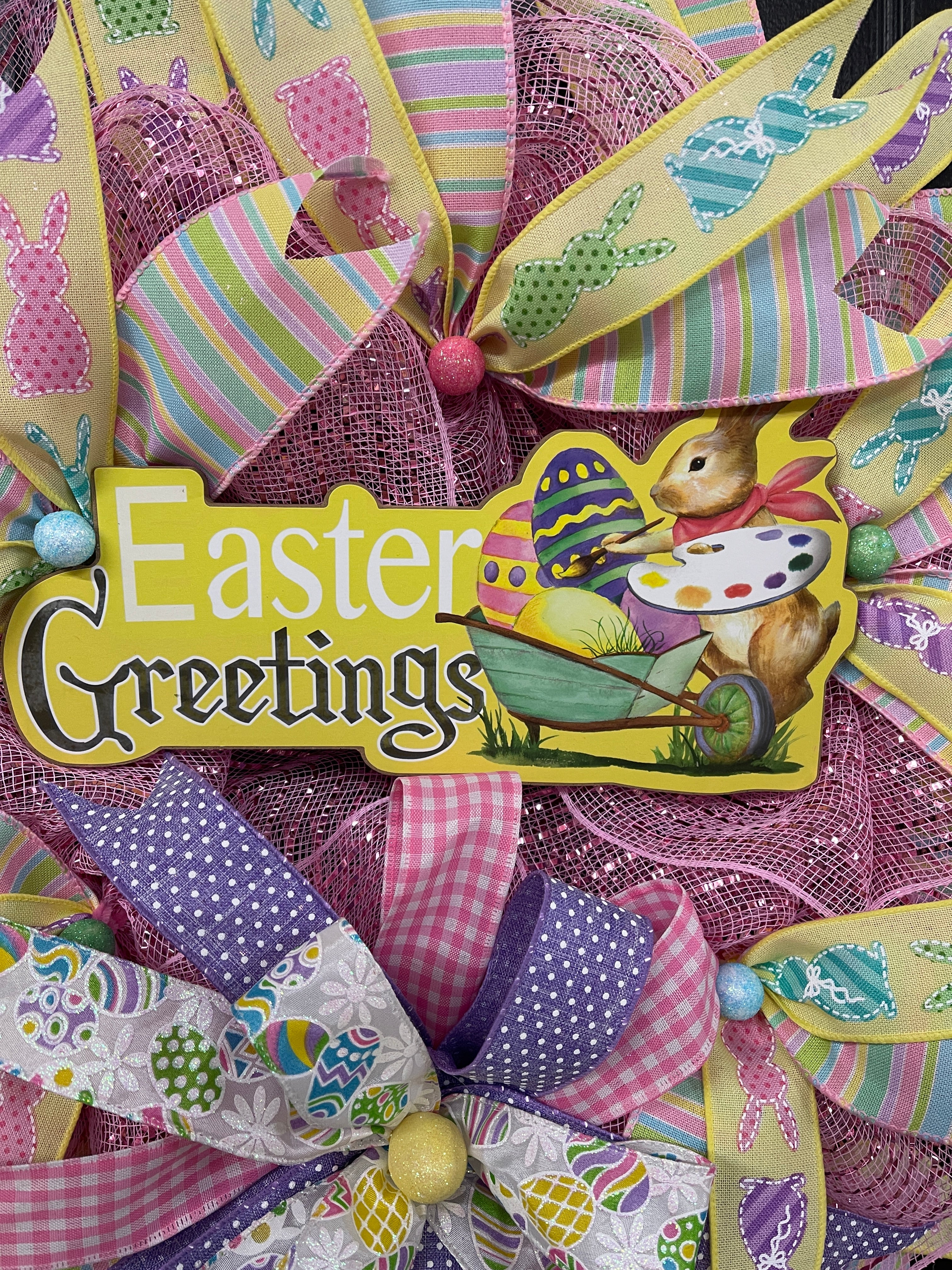 Close up of Easter Greetings Sign with Easter Bunny Holding a Paint Palette painting Easter Eggs in a Wheelbarrow