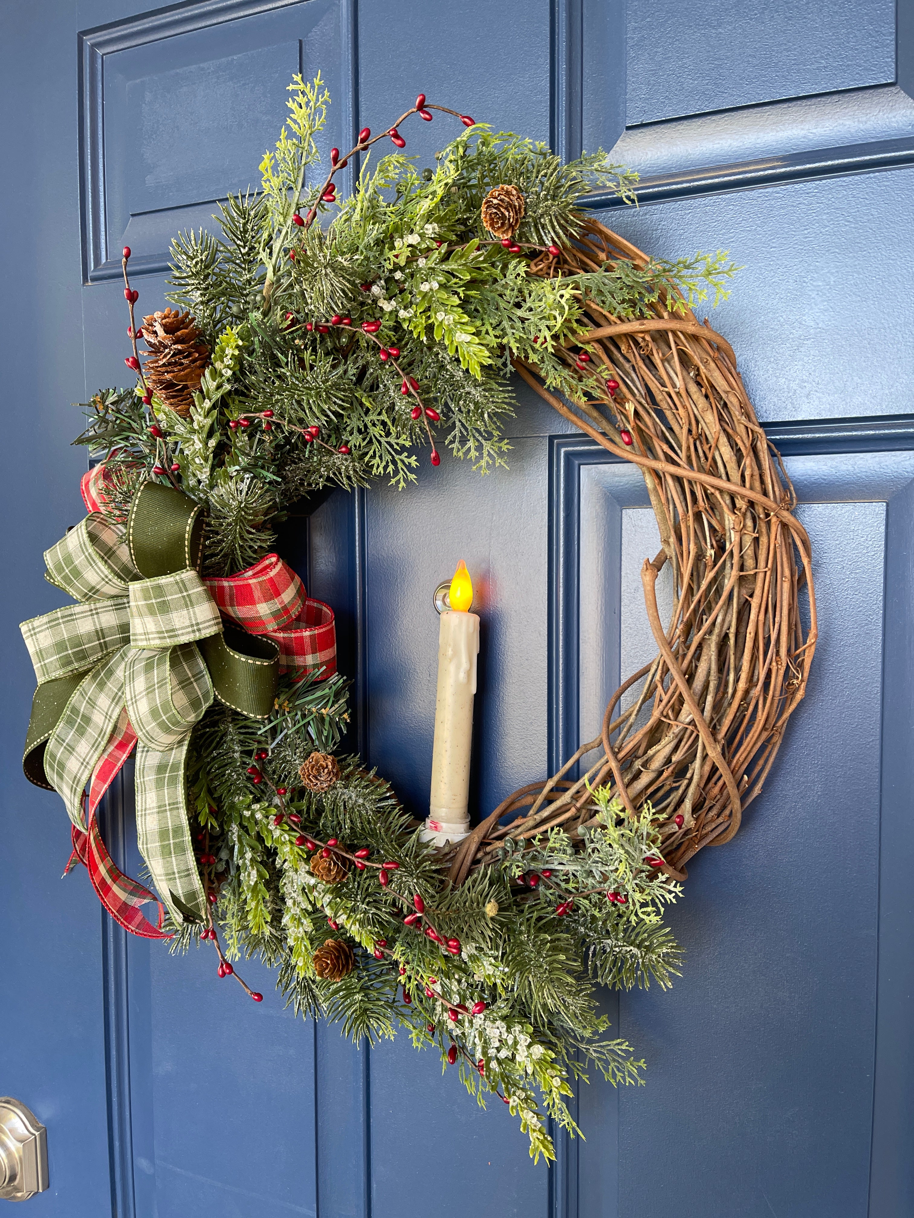Right Side View of Christmas Grapevine Wreath with Artificial Pine Branches, Red Berries, Pinecones, with a Red, White and Green Bow and an Artificial Battery Operated Candle in the Center on a Blue Door