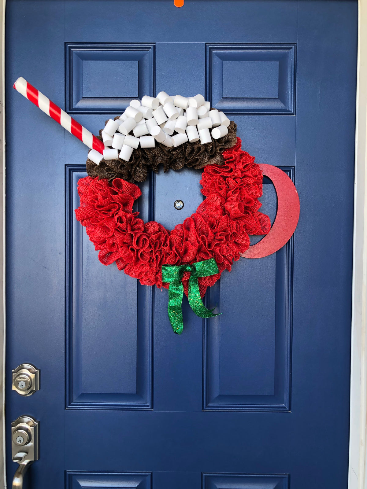 Red Burlap Wreath in the Shape of a Cup of Hot Cocoa with White Foam Marshmallows, a peppermint stick straw and a red wood handle on the cup with a green bow on a blue door. 