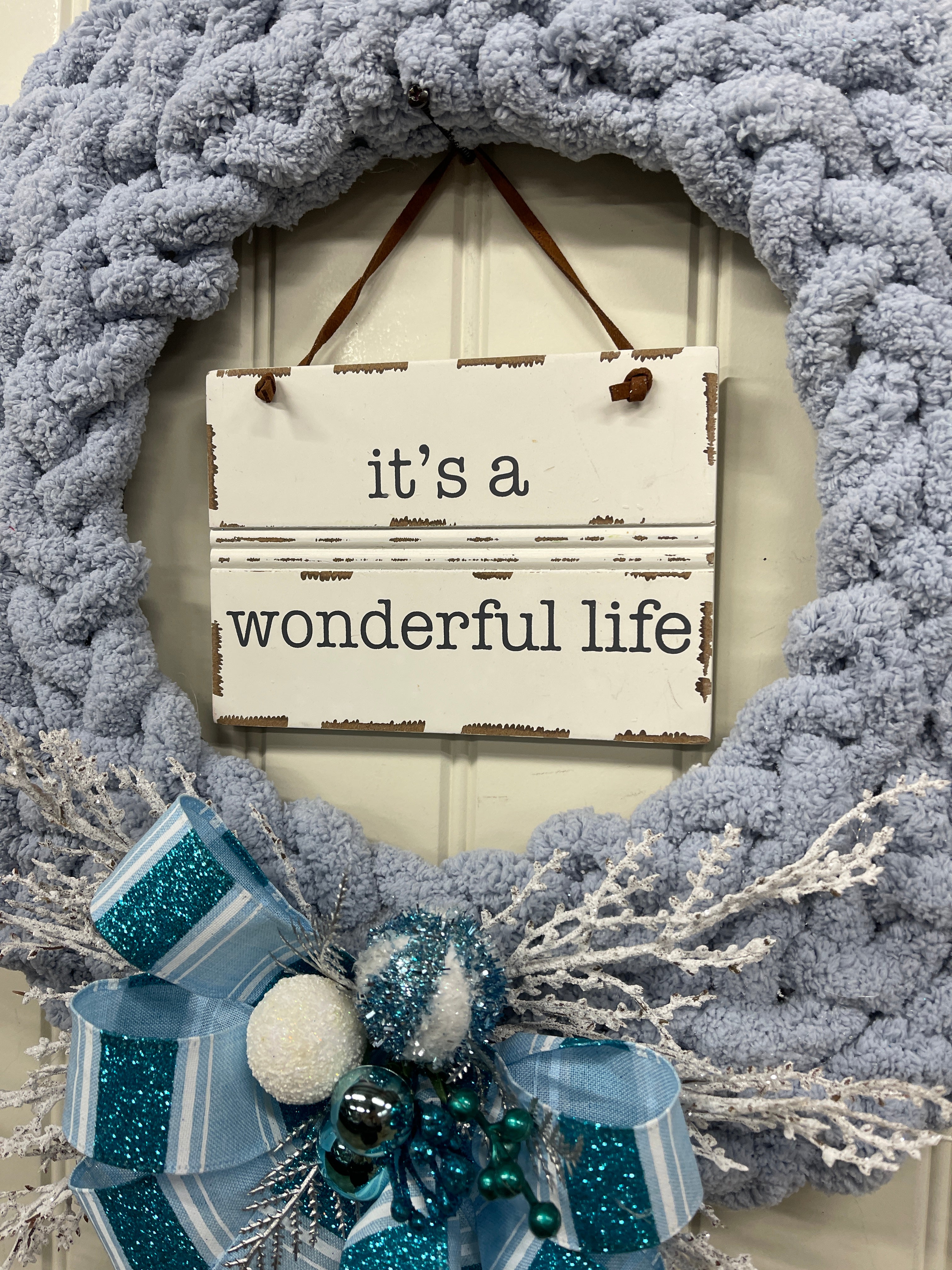 Close up detail of It's a wonderful life sign hanging in the middle of the wreath with a blue and white bow and springs of white frosted pine with blue and white Christmas balls. 