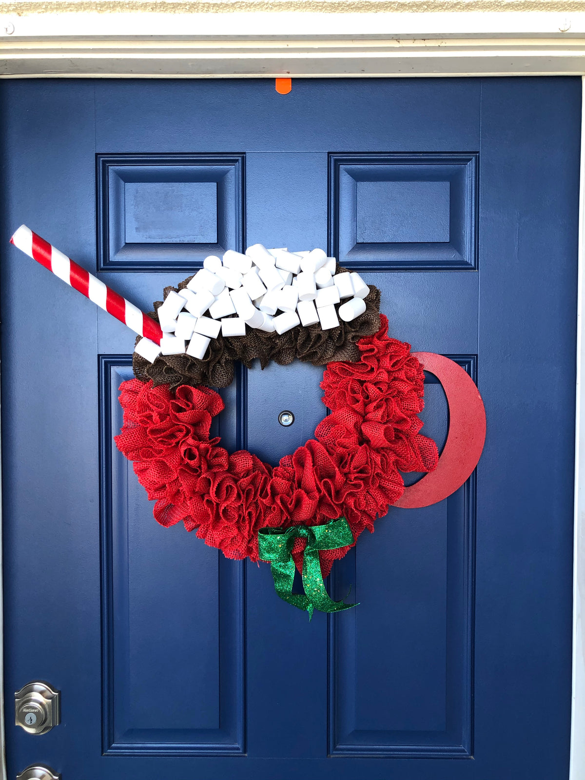 Red Burlap Wreath in the Shape of a Cup of Hot Cocoa with White Foam Marshmallows, a peppermint stick straw and a red wood handle on the cup with a green bow on a blue door. 