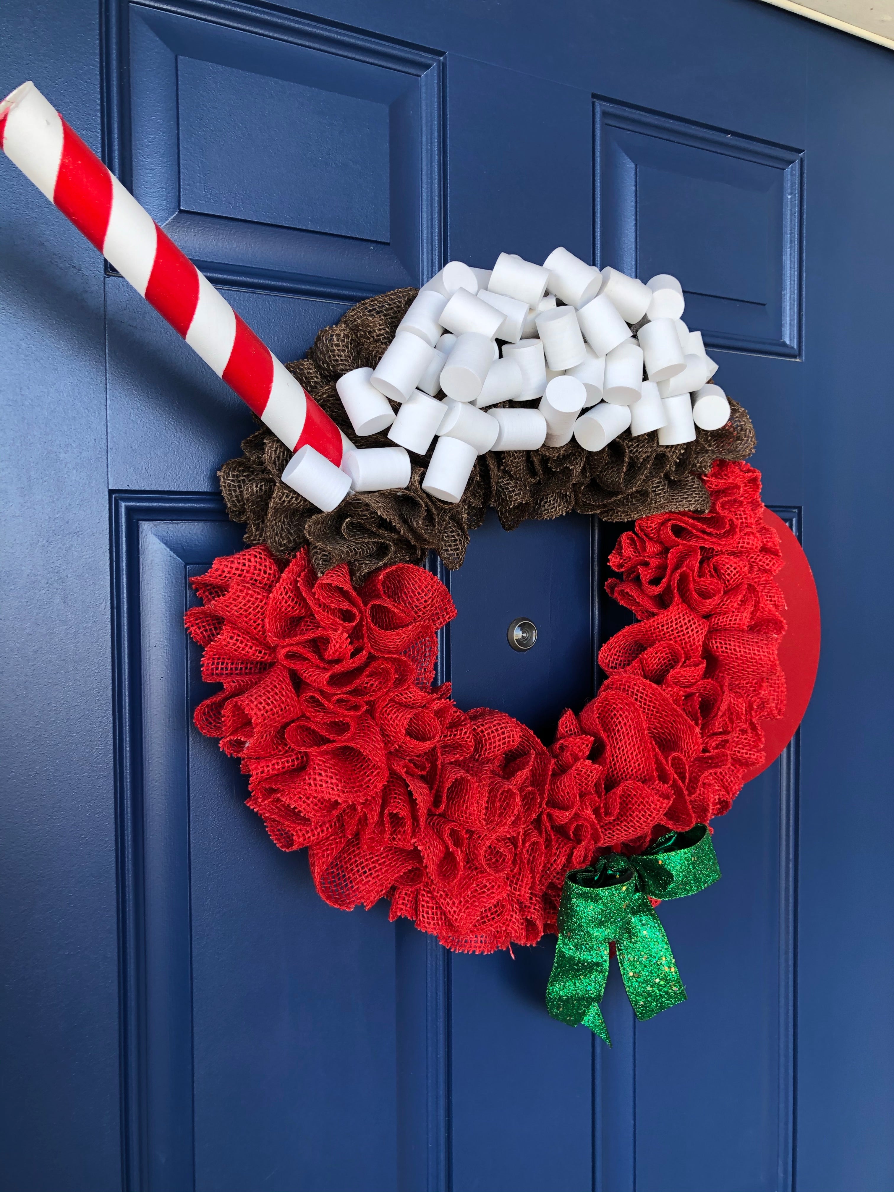 Left Side View of Red Burlap Wreath in the Shape of a Cup of Hot Cocoa with White Foam Marshmallows, a peppermint stick straw and a red wood handle on the cup with a green bow on a blue door. 