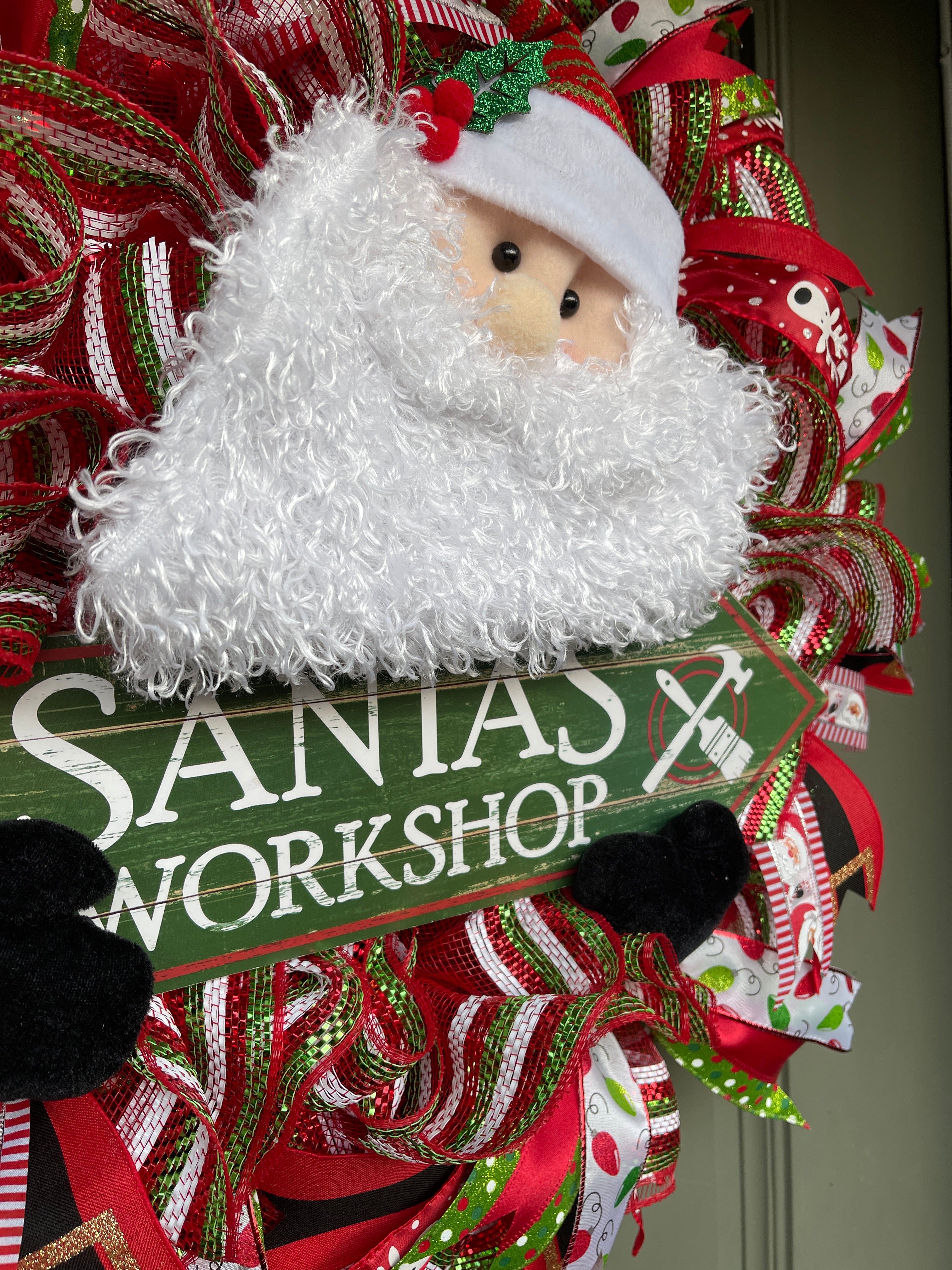 Close Up of Santa Plush wearing a red and Green Striped Hat with Holly holding a green sign with Santa's Workshop in White. 