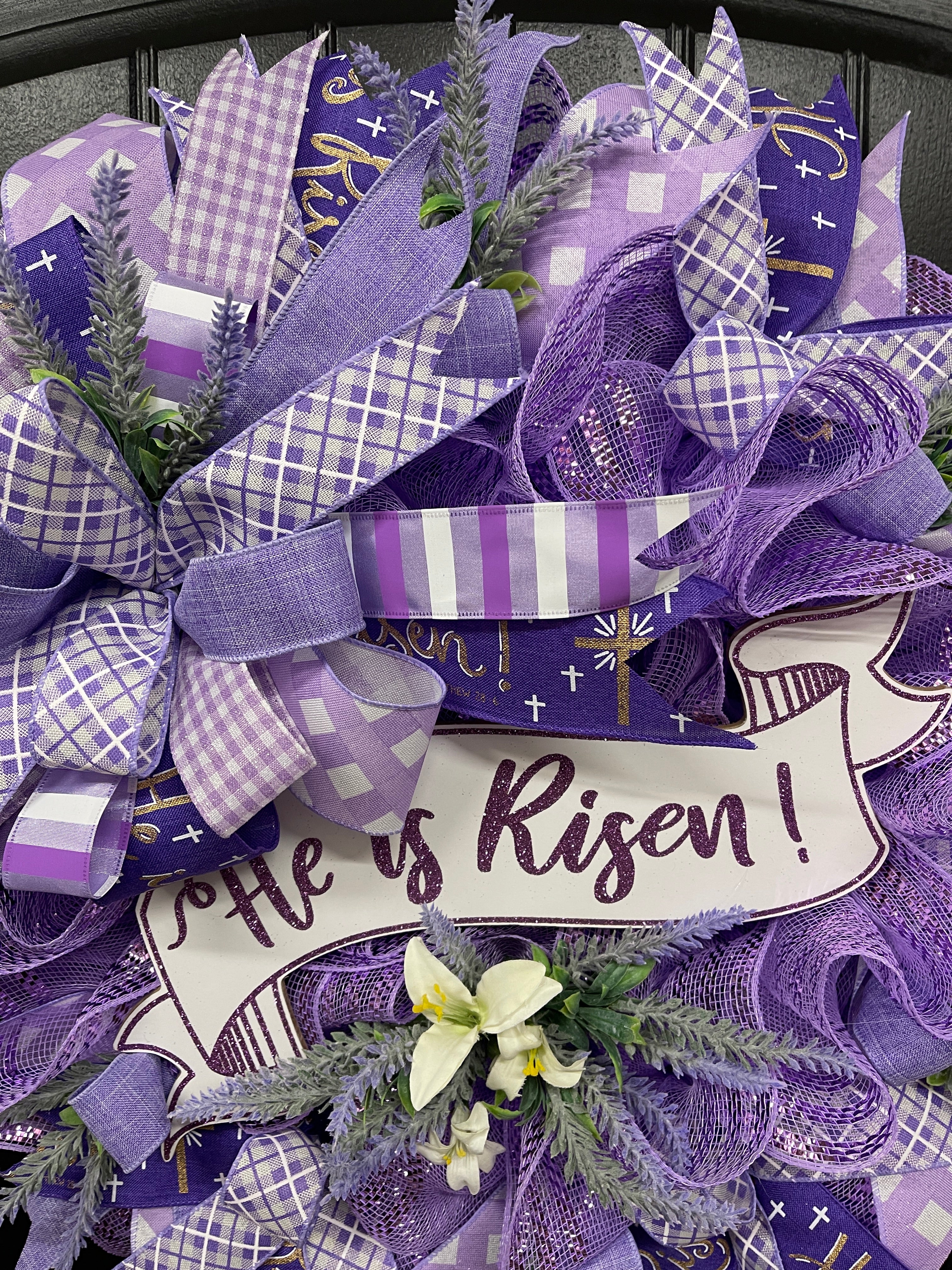 Close Up of Lavender Colored Deco Mesh Wreath Featuring a Purple and White Bow, He is Risen Banner Sign and Artificial florals of Lavender and White Lilies. 