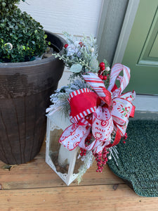 Left Side View of Red and White Candy Cane and Peppermint Lantern Swag with Evergreen Florals and Red Berries on a Wood Porch next to a Potted Plant. 