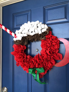 Right Side View of Red Burlap Wreath in the Shape of a Cup of Hot Cocoa with White Foam Marshmallows, a peppermint stick straw and a red wood handle on the cup with a green bow on a blue door. 