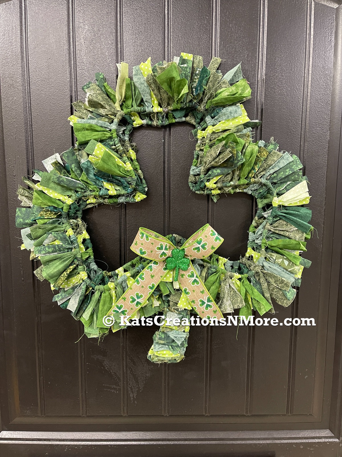 Various Light and Dark Shades of Green Cotton Fabric tied onto a Clover Shaped Frame topped off with a beige and clover ribbon and a glittered green shamrock on a black door. 