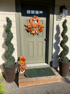 Far Away View of Fall Leaves of Brown, Yellow and Orange, with Orange and Yellow Mums, Pumpkins , Cinnamon Sticks, Red Berries and Pine Cones on a White Lantern on the Porch with a Green Front Door with A Letter S Wreath , along with 2 topiary trees in brown pots. 