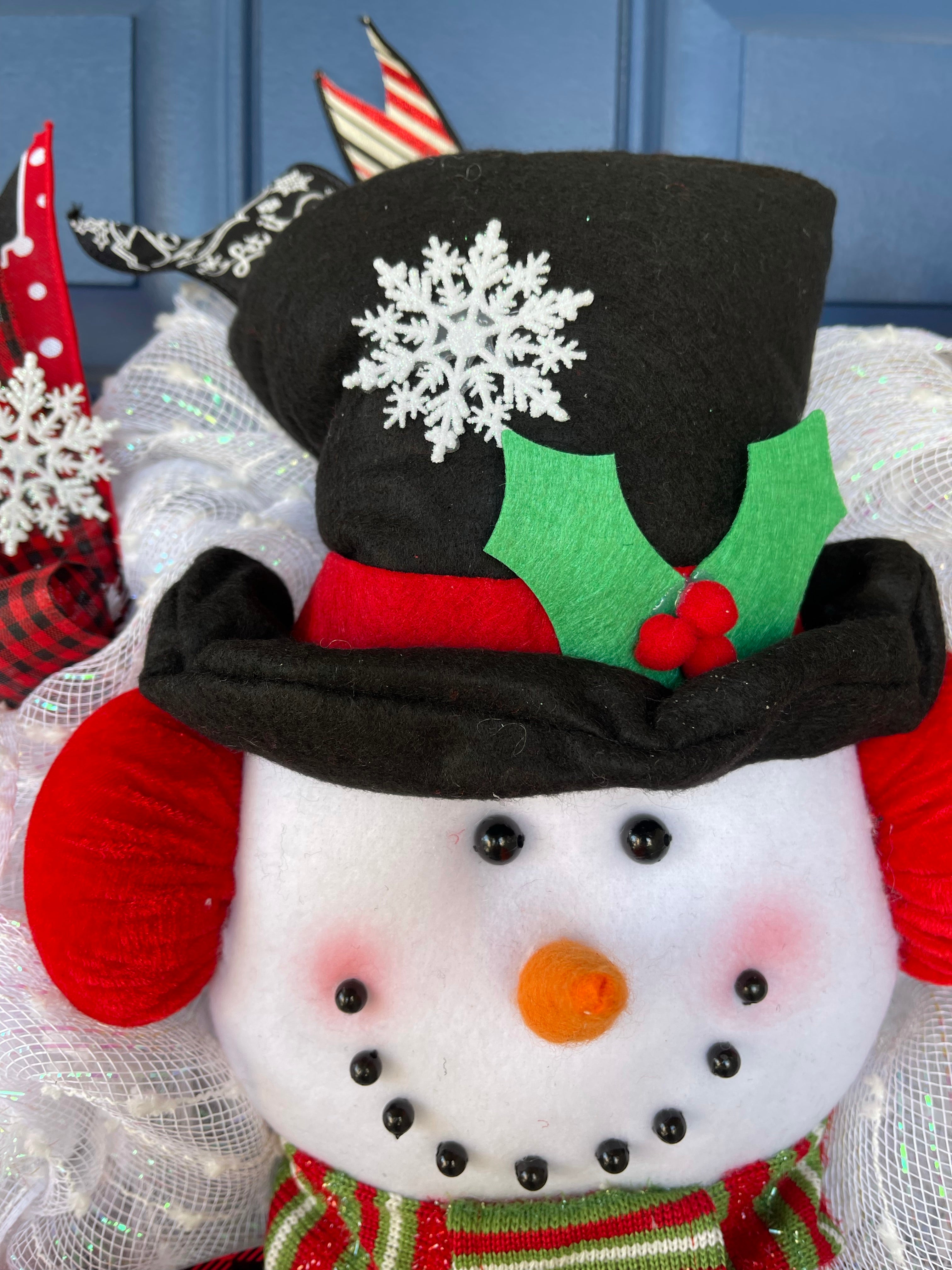 Close Up Detail of Plush Snowman Face with A Carrot Nose, wearing a Black Top Hat with a Snowflake, and Red Brim with Holly Berries, wearing Red Ear Muffs and a Green, White and Red Scarf