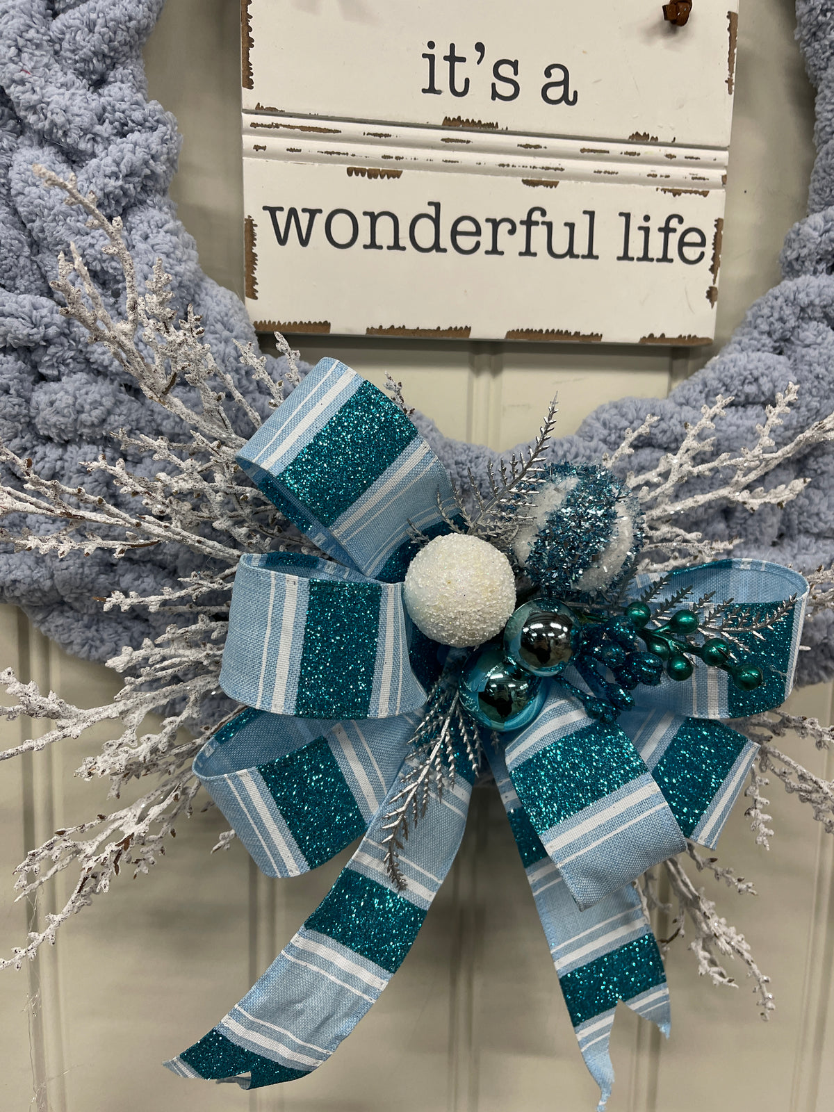 Close Up Detail of Blue and White Bow at the Bottom of the wreath with blue and white balls, silver pine sprigs and white frosted branches. 